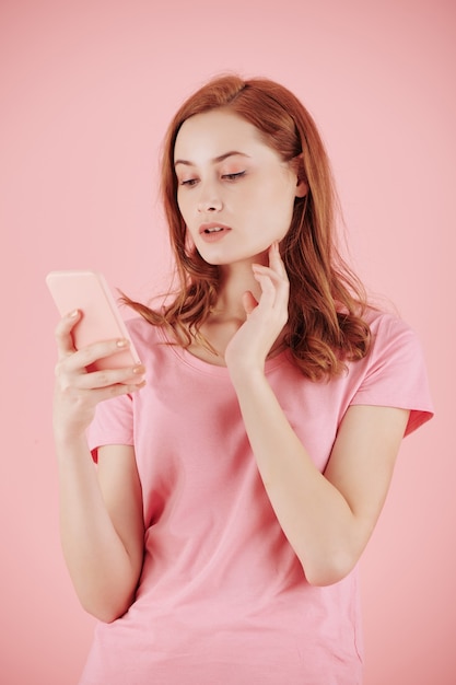 Belle jeune femme aux cheveux rouges lisant des messages texte sur smartphone, isolé sur rose