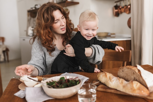 Belle jeune femme aux cheveux rouges en pull tricoté assis à la table avec de la nourriture nourrissant rêveusement son petit fils souriant