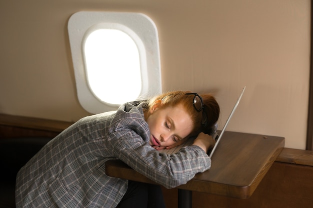 Belle jeune femme aux cheveux rouges vole dans un avion de classe affaires et dort sur un ordinateur portable