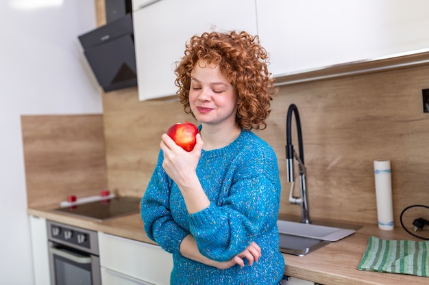 Belle jeune femme aux cheveux roux bouclés manger une pomme rouge juteuse en se tenant debout dans sa cuisine à la maison. Apport quotidien de vitamines aux fruits, alimentation et alimentation saine