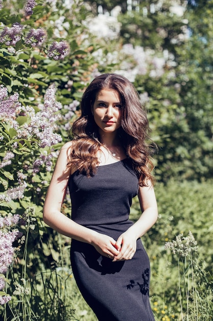 Belle jeune femme aux longs cheveux bouclés brune en robe noire marchant dans le parc d'été