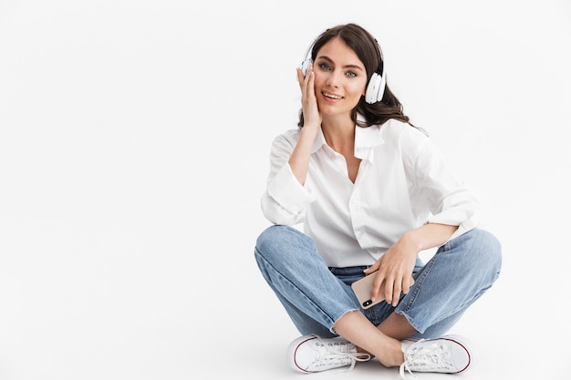 Belle jeune femme aux longs cheveux bouclés brune vêtue d'une chemise blanche assise isolée sur un mur blanc, appréciant d'écouter de la musique avec des écouteurs