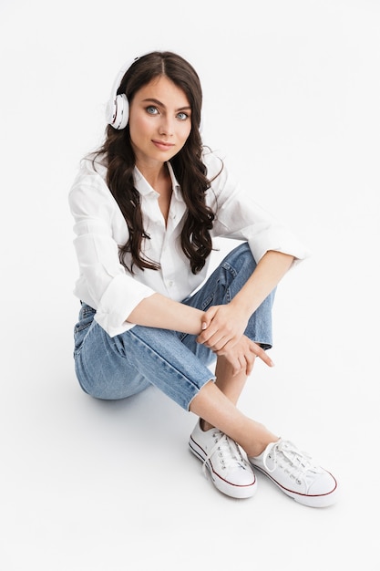 Belle jeune femme aux longs cheveux bouclés brune vêtue d'une chemise blanche assise isolée sur un mur blanc, appréciant d'écouter de la musique avec des écouteurs