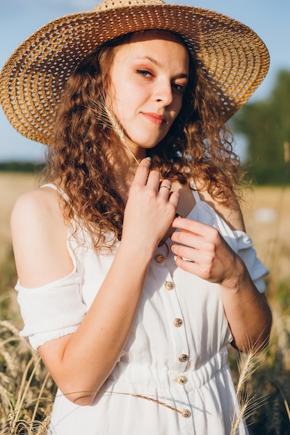 Belle jeune femme aux longs cheveux bouclés pose dans un champ de blé en été au coucher du soleil