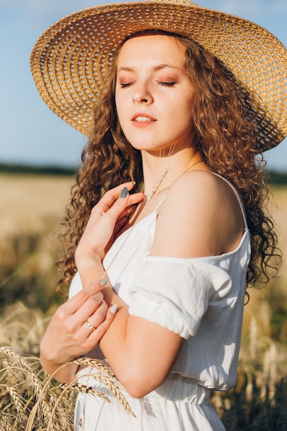 Belle jeune femme aux longs cheveux bouclés pose dans un champ de blé en été au coucher du soleil