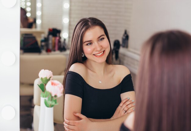 Belle jeune femme aux longs cheveux brune en bonne santé en regardant le miroir dans un salon de coiffure