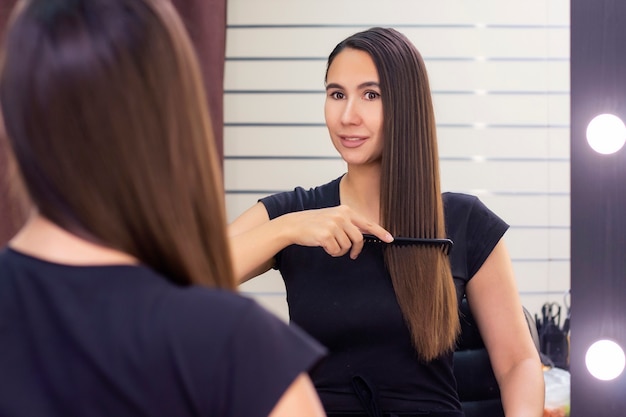 Belle jeune femme aux longs cheveux bruns lisses prend soin de ses cheveux. se peigner les cheveux et se regarder dans le miroir