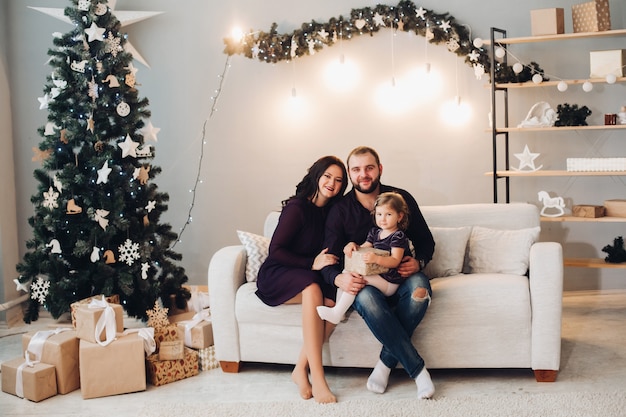 Belle jeune femme aux longs cheveux noirs ondulés en robe, homme fort attrayant avec des cheveux noirs courts en chemise et jeans passent le temps de Noël avec une petite fille