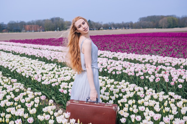 Belle jeune femme aux longs cheveux roux portant en robe blanche debout avec une vieille valise vintage sur champ de tulipes colorées.