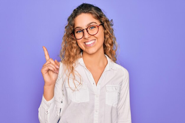 Belle jeune femme aux yeux bleus portant une chemise décontractée et des lunettes sur fond violet montrant et pointant vers le haut avec le doigt numéro un tout en souriant confiant et heureux