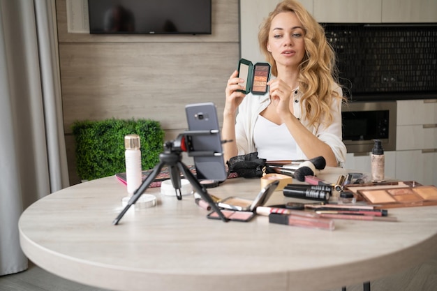 Une belle jeune femme blogue sur le maquillage et les produits de beauté Superbe dame qui enlève un tutoriel de maquillage avec une palette de fards à paupières de maquillage Une fille souriante enseigne le maquillage à l'aide de son téléphone