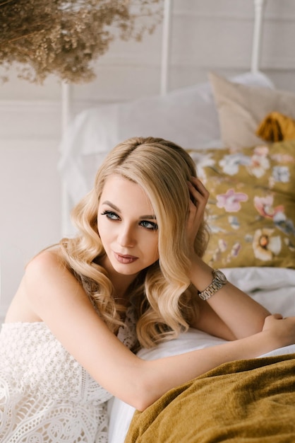 Belle jeune femme blonde aux cheveux bouclés et maquillage dans une robe blanche à l'intérieur lumineux de la maison studio