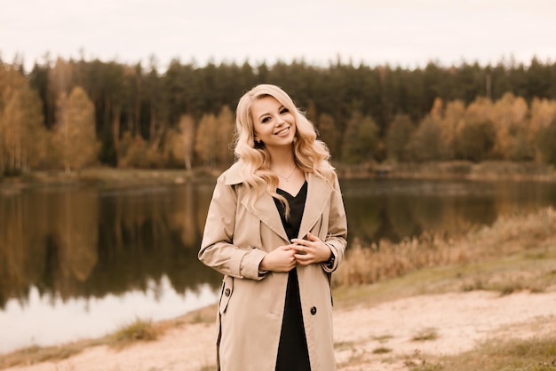 Belle jeune femme blonde aux cheveux bouclés et maquillage dans un trench-coat beige sur le fond du lac d'automne doré