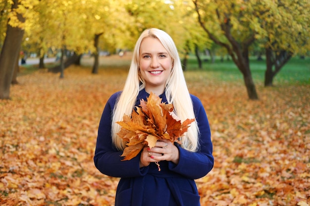 Une belle jeune femme blonde aux cheveux longs dans un manteau bleu et avec un bouquet de feuilles d'automne i