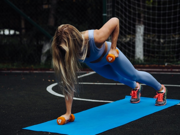 Belle jeune femme blonde aux cheveux longs en vêtements de sport bleu clair faire des exercices avec des cloches en plein air dans l'arrière-cour
