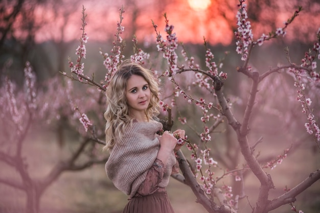 Belle jeune femme blonde bouclée en jupe plissée marron chemisier rose épaules couvertes avec écharpe tricotée, se dresse dans les jardins de coucher de soleil de pêche en fleurs