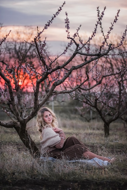 Belle jeune femme blonde bouclée en jupe plissée marron, chemisier rose, épaules couvertes avec écharpe tricotée, se trouve dans les jardins de coucher de soleil de pêche en fleurs