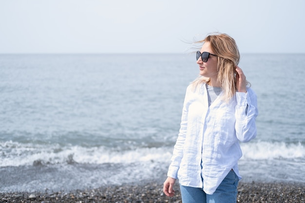 Belle jeune femme blonde en chemise blanche et lunettes de soleil debout au bord de la mer.