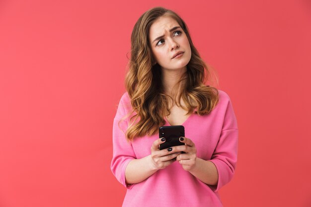 Belle jeune femme blonde confuse debout isolée sur un mur rose, tenant un téléphone portable