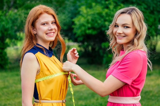 Belle et jeune femme blonde couturière couturière mesure le corps avec un ruban à mesurer de fille norvégienne irlandaise rousse au gingembre dans une robe jaune dans le parc d'été. tailleur et client à l'extérieur