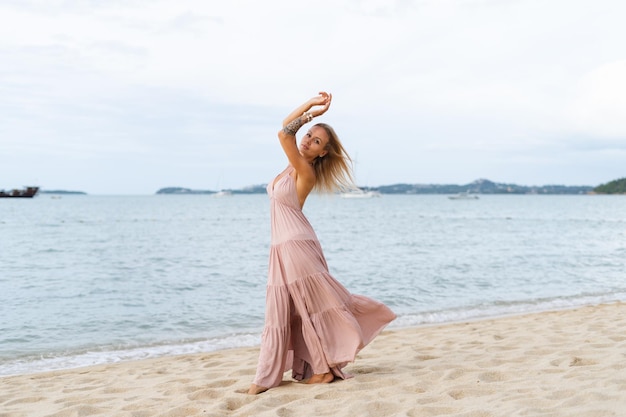 Belle jeune femme blonde dans une longue robe rose sur la plage tendre posant le vent développe la robe cheveux romantique libre