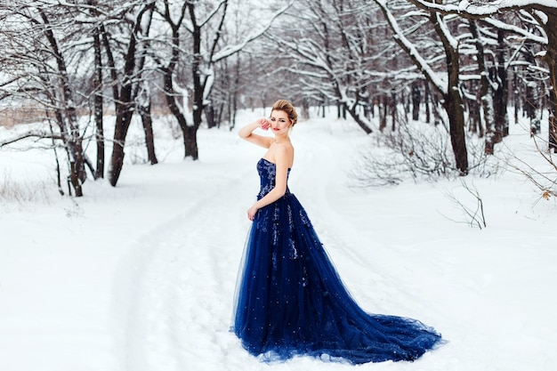 Une belle jeune femme blonde dans une robe bleue luxuriante posant dans un parc d'hiver enneigé