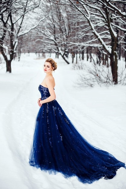 Une belle jeune femme blonde dans une robe bleue luxuriante posant dans un parc d'hiver enneigé