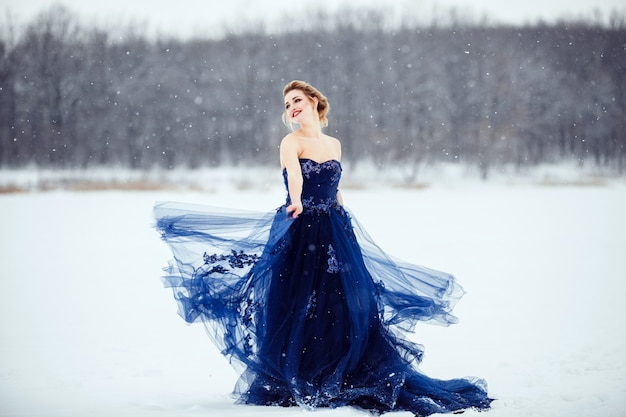 Une belle jeune femme blonde dans une robe bleue luxuriante posant dans un parc d'hiver enneigé