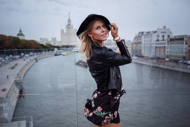 Une belle jeune femme blonde dans une veste en cuir noir, une jolie robe courte et un chapeau se promène dans le parc Zaryadye, Moscou.