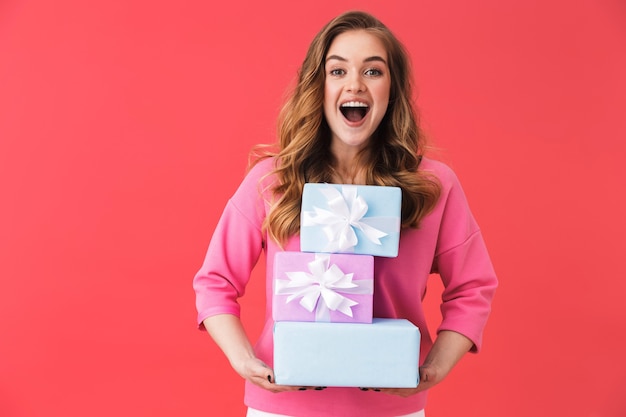 Belle jeune femme blonde debout isolée sur un mur rose, portant une pile de coffrets cadeaux