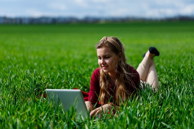 Belle jeune femme blonde est allongée sur l'herbe verte dans le parc avec un ordinateur portable et travaille. Ciel bleu avec des nuages. La fille sourit et profite d'une bonne journée. Travaillez sur la nature par une journée ensoleillée.