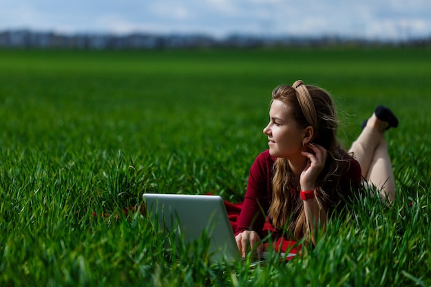 Belle jeune femme blonde est allongée sur l'herbe verte dans le parc avec un ordinateur portable et travaille. Ciel bleu avec des nuages. La fille sourit et profite d'une bonne journée. Travaillez sur la nature par une journée ensoleillée.