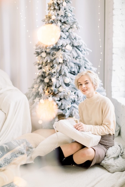 Belle jeune femme blonde est assise sur le sol près de l'arbre de Noël