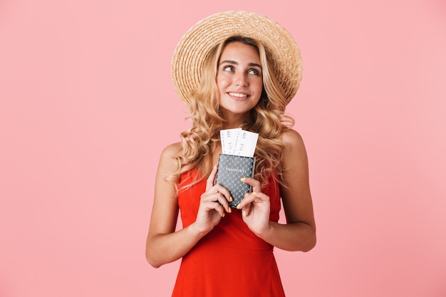 Belle jeune femme blonde heureuse vêtue d'une robe d'été isolée sur un mur rose, montrant un passeport avec des billets d'avion