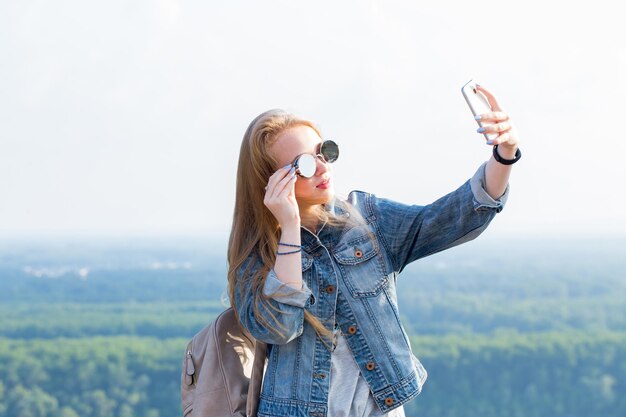 Belle jeune femme blonde à lunettes de soleil prend un selfie contre le paysage