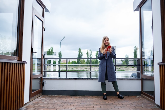 Belle jeune femme blonde en manteau gris parlant au téléphone par une froide journée d'automne. Fille avec téléphone sur la terrasse surplombant la rivière