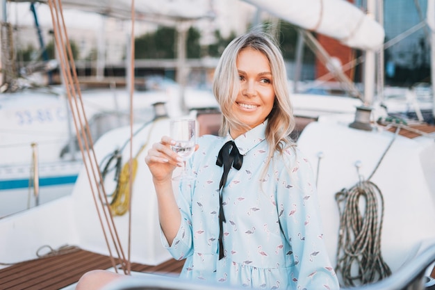 Belle jeune femme blonde en robe bleue avec un verre de soda sur le bateau à l'embarcadère au coucher du soleil