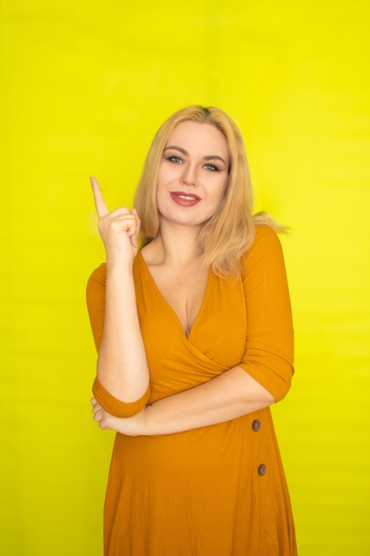 Belle jeune femme blonde en robe jaune foncé posant sur un mur jaune à l'intérieur