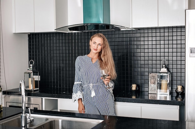 Belle jeune femme blonde en robe rayée pose dans la cuisine avec un verre de vin à la main