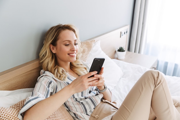 Belle jeune femme blonde se détendre dans son lit à la maison, à l'aide d'un téléphone portable