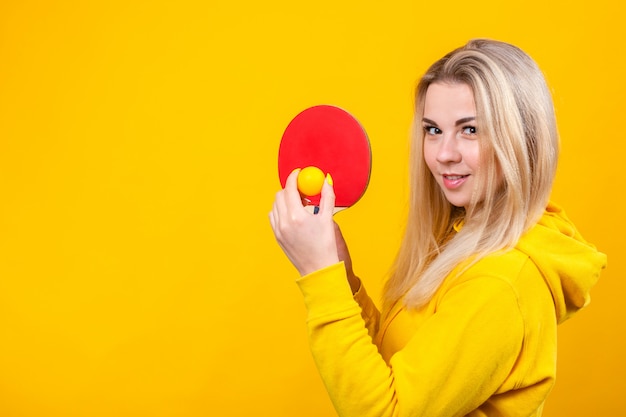 Belle jeune femme blonde séduisante dans des vêtements sportifs décontractés jaunes jouer au ping-pong, tenant une balle et une raquette.