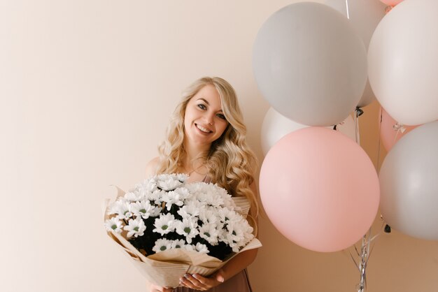 Belle jeune femme blonde souriante avec des ballons blancs gris et roses et des fleurs de marguerites blanches ou de chrysanthèmes sur un mur lumineux, espace copie