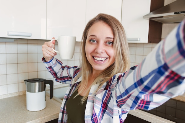 Une belle jeune femme blonde souriante caucasienne dans la chemise à carreaux avec une tasse de café blanc