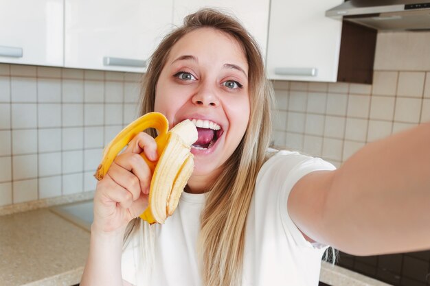 Une belle jeune femme blonde souriante caucasienne dans un tshirt blanc mange une banane et prend un selfie