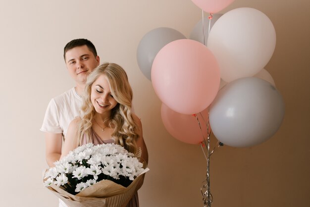 Belle jeune femme blonde souriante et mec avec des ballons et des fleurs de marguerites blanches ou de chrysanthèmes sur un mur léger