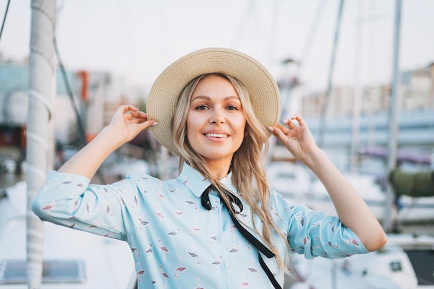 Belle jeune femme blonde souriante en robe bleue et chapeau de paille à l'embarcadère au coucher du soleil