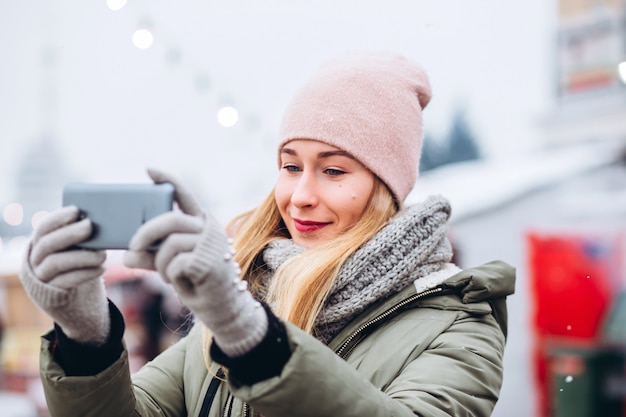 Une belle jeune femme blonde souriante à un téléphone pour une photo sur le marché de Noël