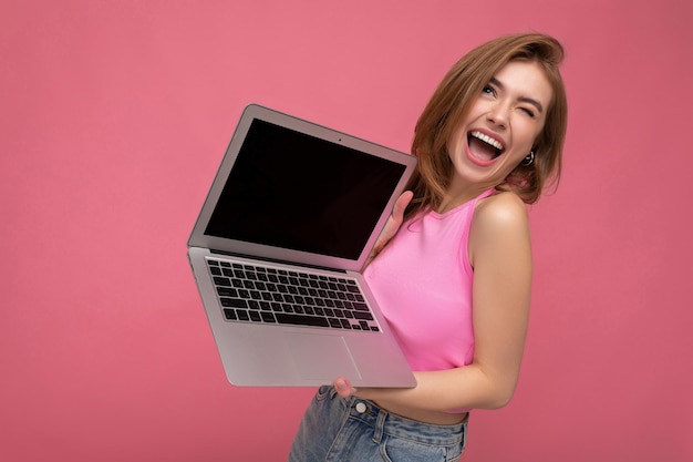 Belle jeune femme blonde souriante tenant un ordinateur portable portant un haut court rose en levant