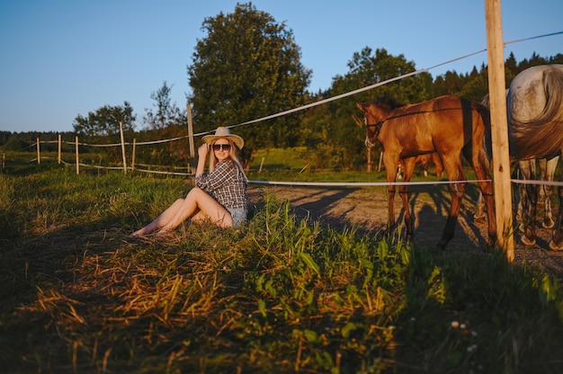 Belle jeune femme blonde vêtue de style safari en chapeau et chemise à carreaux posant avec des pur-sang