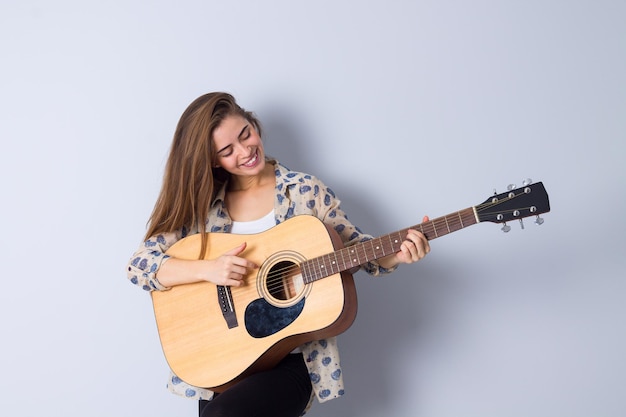 Belle jeune femme en blouse beige et pantalon noir jouant de la guitare sur fond gris en studio